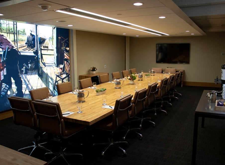 The Belz Boardroom at the Crawford Hotel in Denver, Colorado. A long boardroom table, coffee refreshment station, large television and decorative mural make up the room.