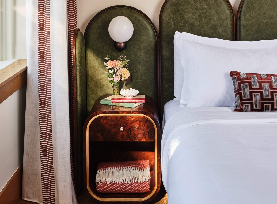 The bedside area of the Deluxe Classic Guest room at the Crawford Hotel in Denver, Colorado. The area has a small table with an extra blanket, fresh cut flowers and a lamp.