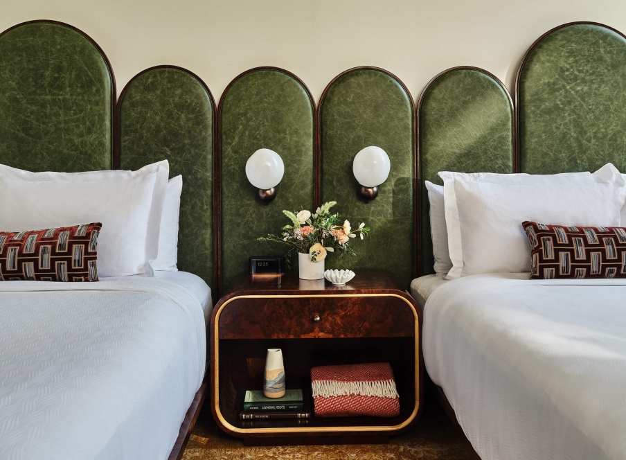 Two queen beds in the Deluxe Classic Guest Room at the Crawford Hotel in Denver, Colorado. A side table is placed between them with lamps, fresh cut flowers, alarm clock and extra blankets.