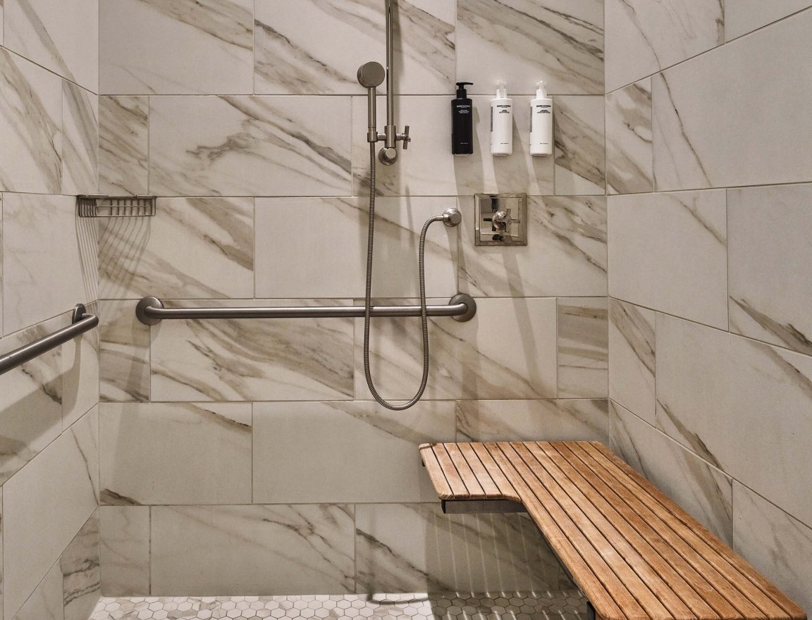 The accessible shower in the Premium Classic ADA room at the Crawford Hotel in Denver, Colorado. The shower area is wide, has a seat near the hand-held shower head with hand bars on the walls.