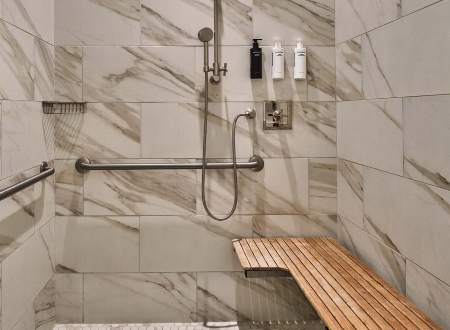 The accessible shower in the Premium Classic ADA room at the Crawford Hotel in Denver, Colorado. The shower area is wide, has a seat near the hand-held shower head with hand bars on the walls.