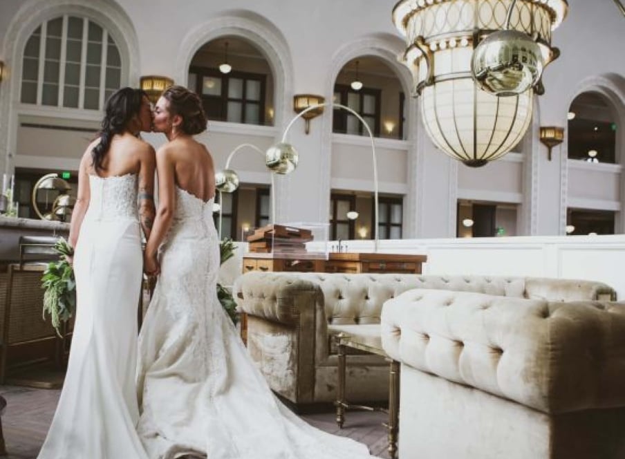 Two brides in white wedding gowns kiss in the lobby of Union Station in Denver, Colorado.
