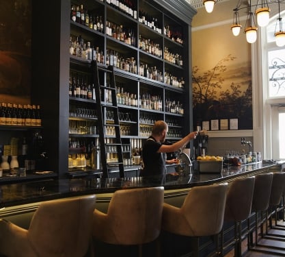 Tall shelving of assorted liquors behind the bar at Ultreia at Union Station in Denver, Colorado. A bar tender prepares drinks.