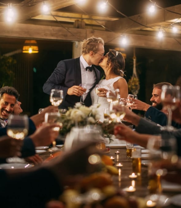 A bride and groom kiss at the head of a table while wedding guests cheers.