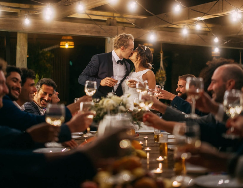 A bride and groom kiss at the head of a table while wedding guests cheers.