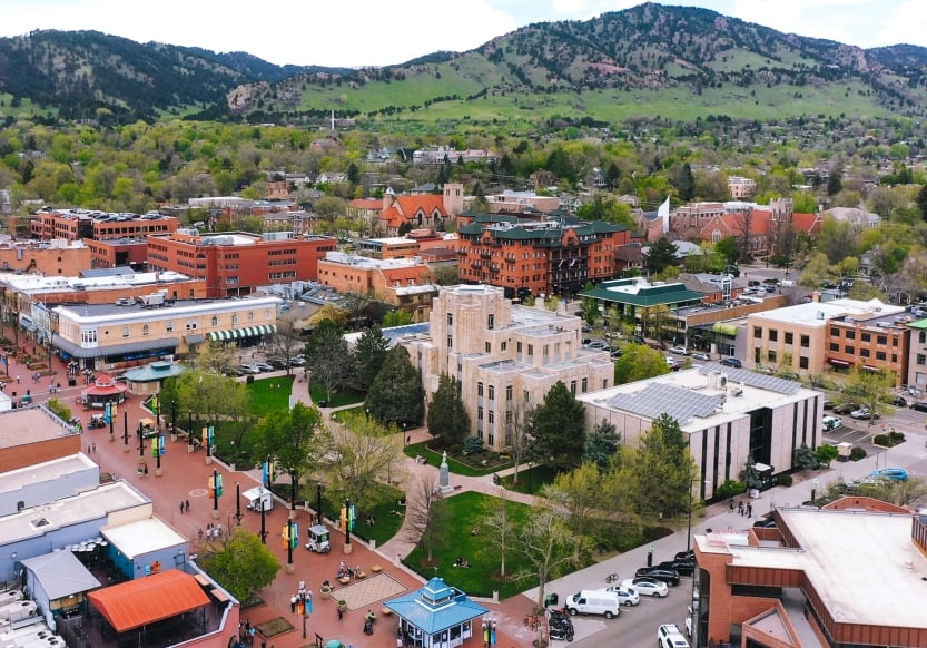 Aerial view of Boulder, Colorado with colorful buildings and streets below, a green mountain range in the background.