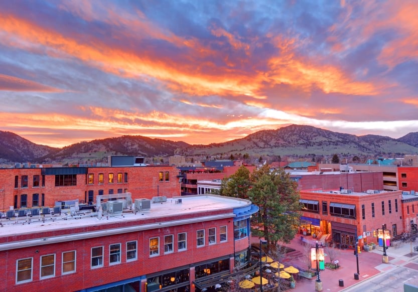 Two-story buildings with patios underneath an orange and purple sunset, a green mountain range in the background.