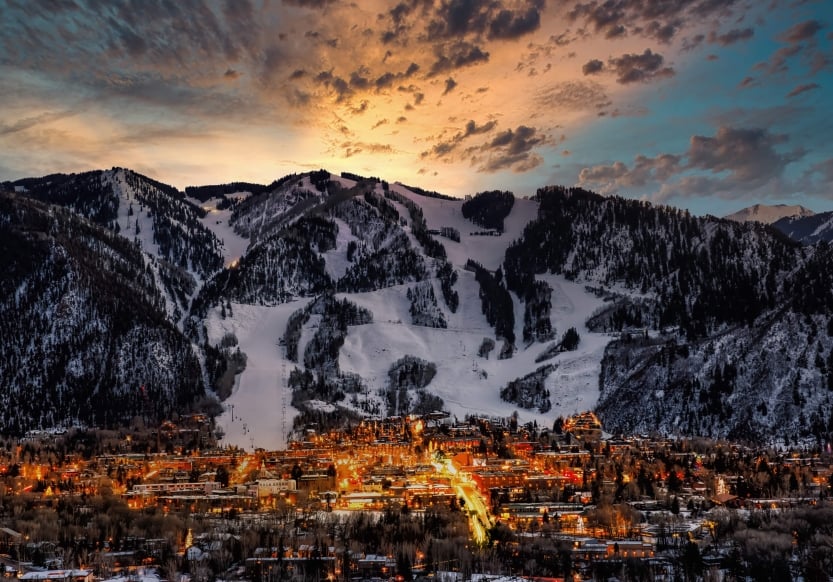 A large snowy mountain towering over a lit-up town at dusk with a sunset in the background.