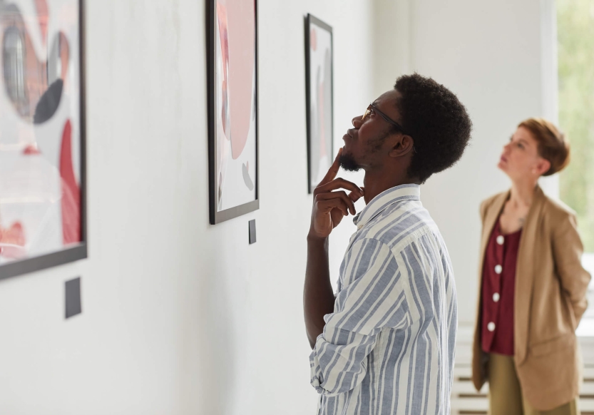 A man thoughtfully looking at art in a gallery, a woman in the background admires the art.