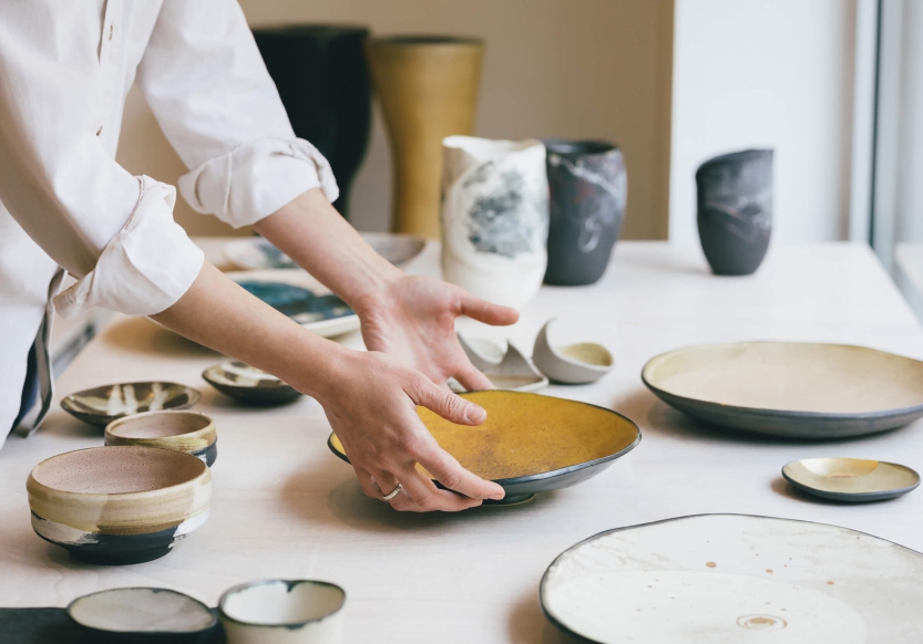 A potter places a hand made plate on a table amongst other pottery pieces.