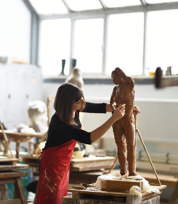 A sculptor in a bright studio moulds a figure in clay.