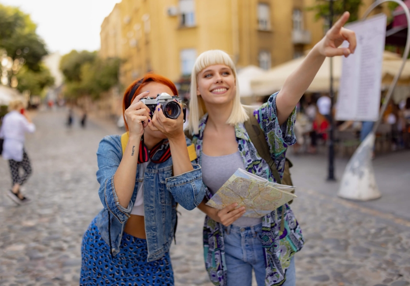 Two women tourists, one taking a picture with a film camera. The other is holding a map while smiling and pointing into the distance.