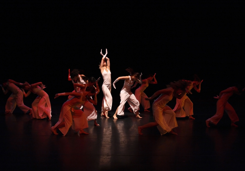 A dramatically lit dance performance. One dancer in the middle is illuminated while others performing around her are in the shadows.