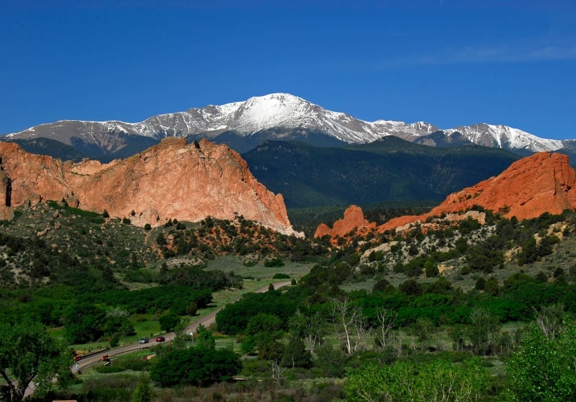 Cars on a road surrounded by green woodlands, driving toward vast, red rock formations with snow capped mountains in the background.