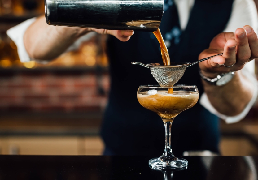 A bar tender pours an espresso martini from a shaker through a strainer into a martini glass.