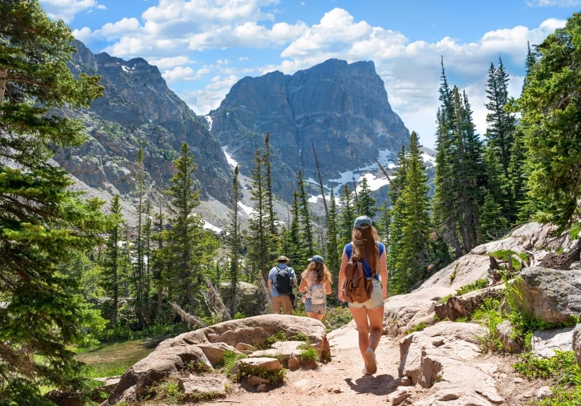 Three people in summer attire hiking into a rocky valley. There are a few ever-green trees in the foreground and a towering rocky mountain range in the background.