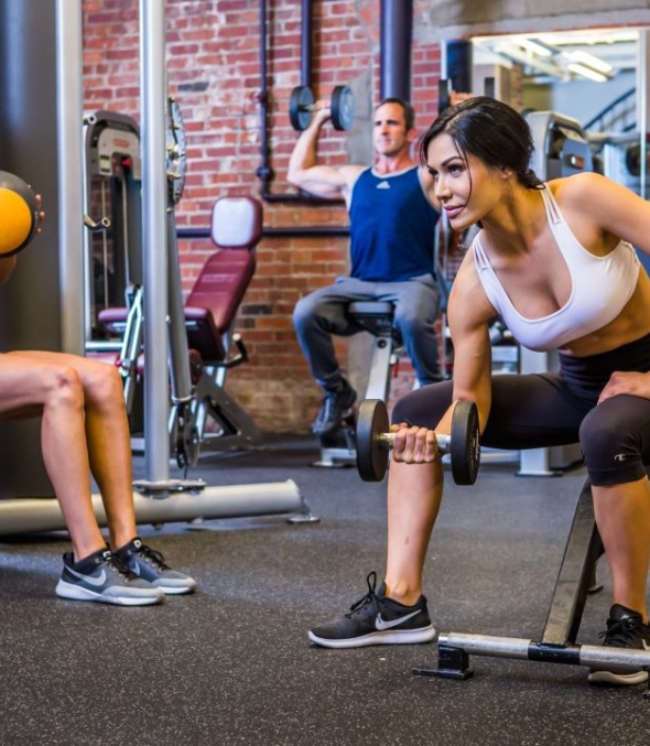 Inside a gym one woman lifts a barbell while another holds a medicine ball and sits against a wall. A man presses weights in the background.