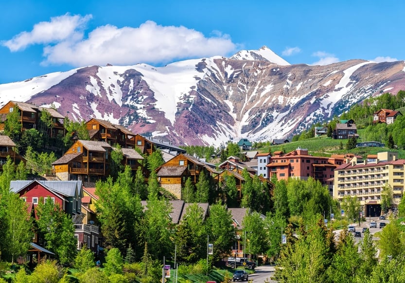 A few buildings scattered on a lush hillside with snow-capped mountains in the background on a sunny day.